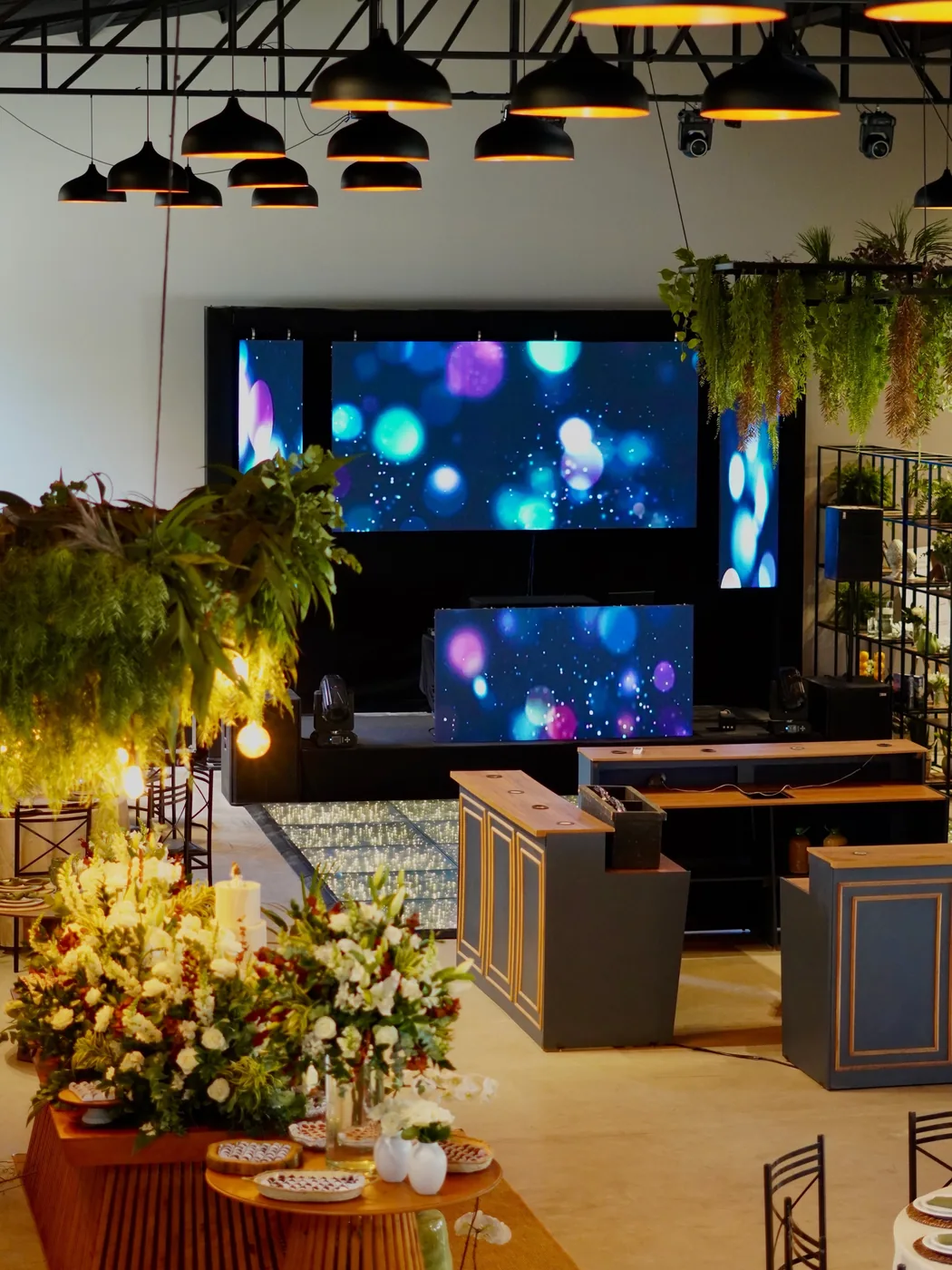 Salão do Recanto das Palmeiras com telão, iluminação pendente e decoração de flores e folhagens